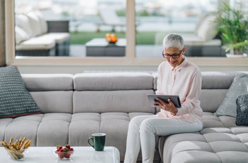 A woman with white hair checks her tablet while seated on a gray couch in a contemporary home.