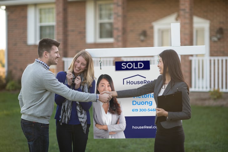 A homebuyer shakes the hand of his real estate agent in front of the sold sign on the lawn of their new brick home. Between them, a woman standing next to the sign cheers the sale.