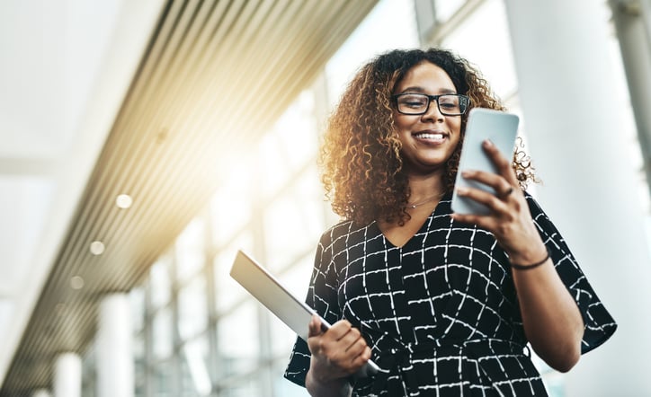 A smiling woman holding a tablet in a brightly lit atrium checks her smartphone.