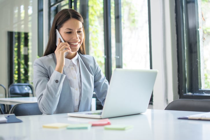 A smiling woman sitting in a well-lot office talks on her smartphone while checking her laptop.