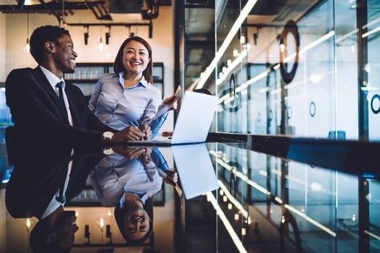 Two young professionals are laughing and smiling in the office after seeing positive changes and results on their laptop.