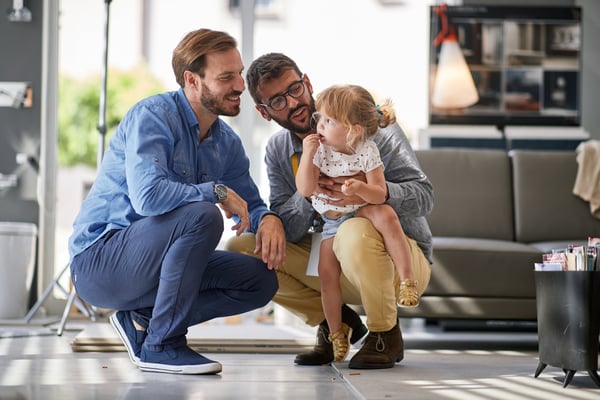 Two young fathers are bent down, playing and laughing with their young daughter in their new home.