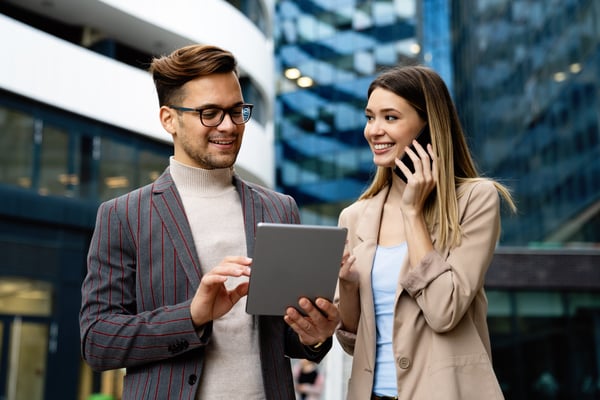 Two young professionals are excited to see a new lead come in. The male is reviewing information on a tablet while the female reaches out to the lead on her cell phone.