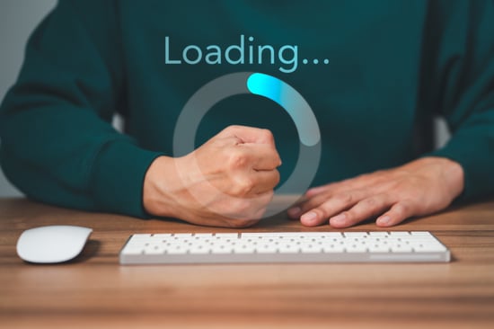 A man is shown sitting at his desk with his fist balled in frustration because his loading time is slow and repetitive.