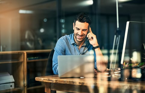 A male real estate agent talks to a client on the phone while checking listing details on his laptop.