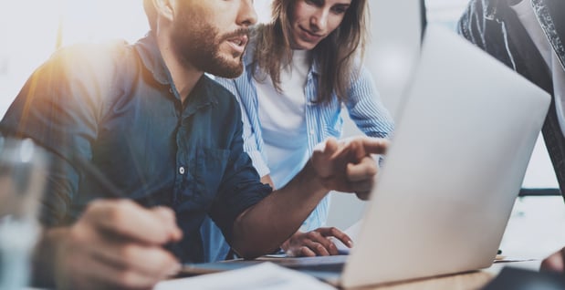 A male agent points at details on his laptop while a female colleague looks over her shoulder in the background.